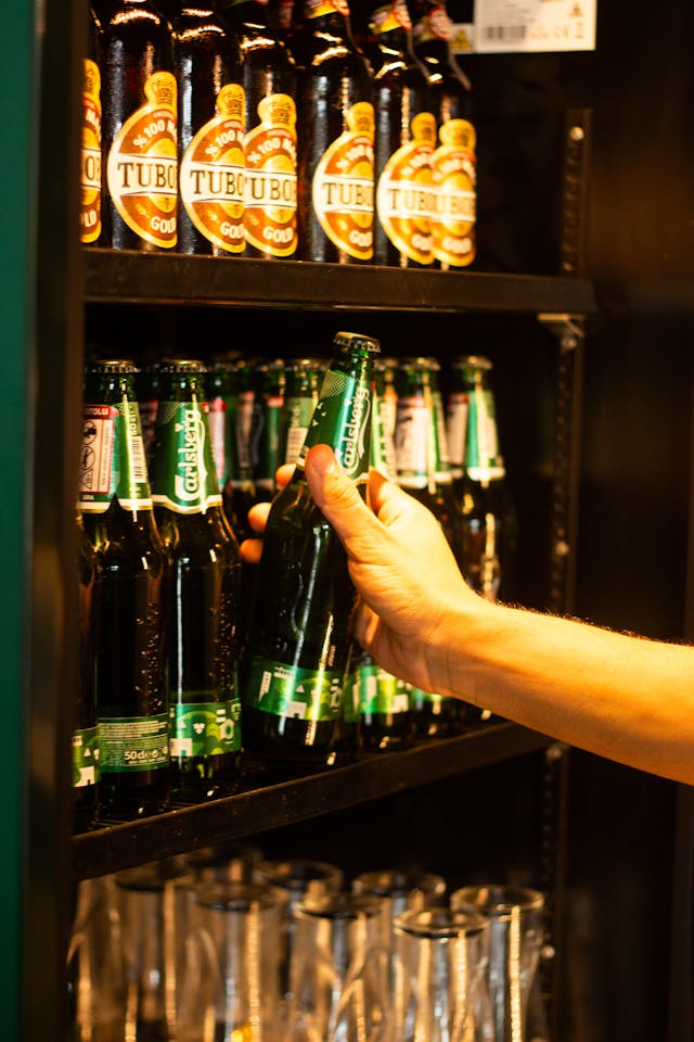 hand retrieving beer bottle from bar fridge filled with beer bottles and cans