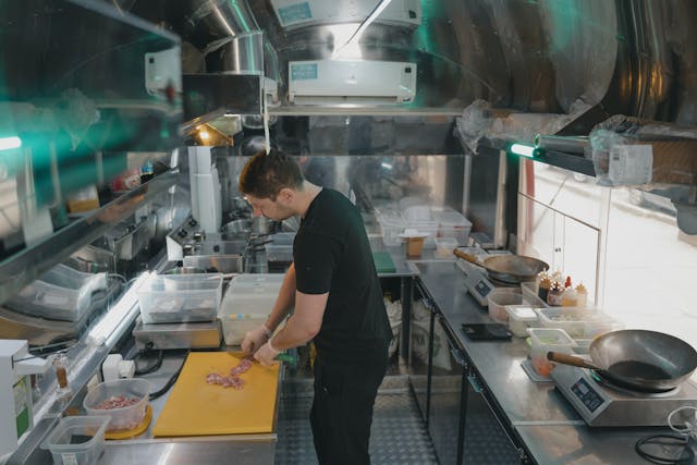 man in small catering kitchen chopping meat on a yellow chopping board